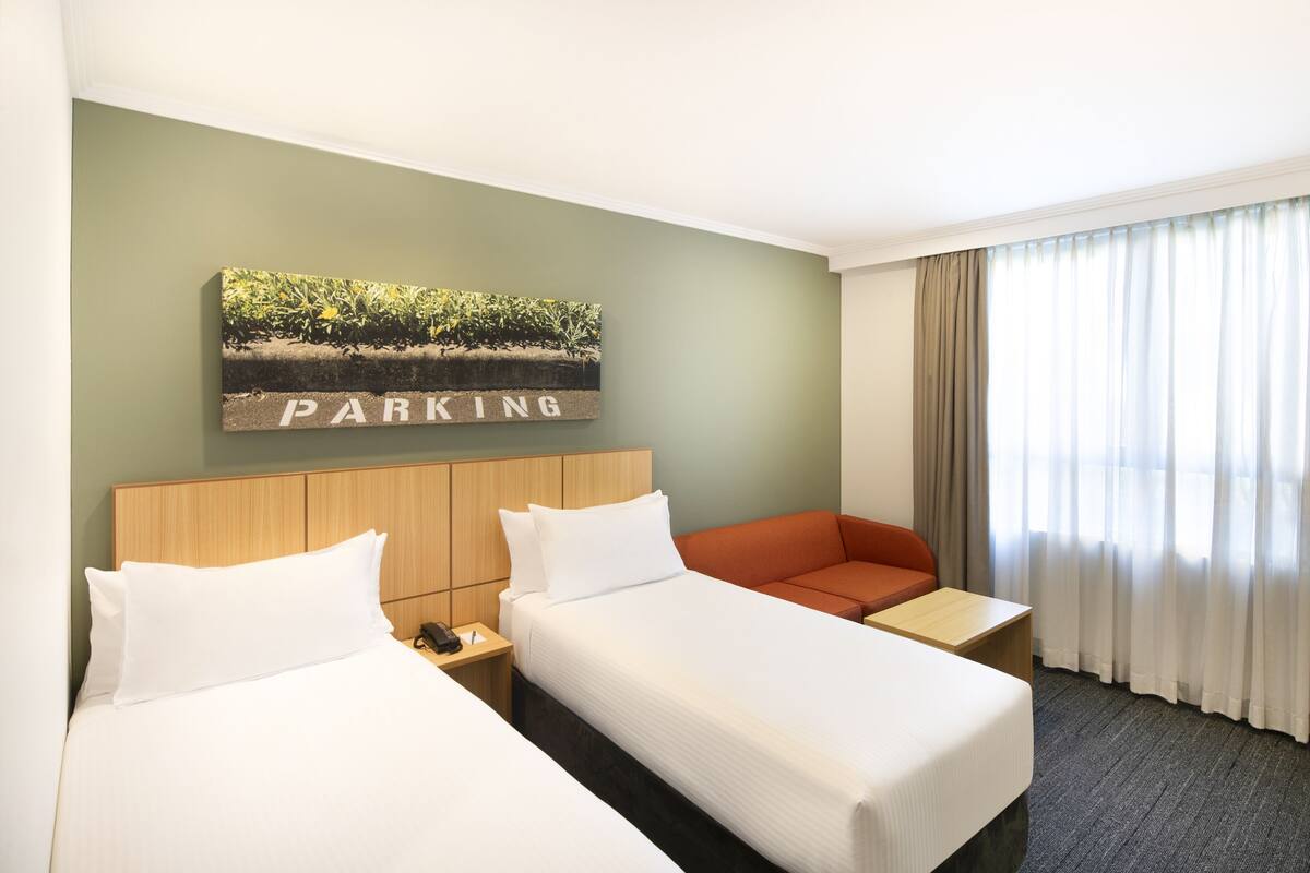 Een hotelkamer met twee bedden, een houten hoofdbord, een schilderij van een landschap, uitzicht op een meer door het raam en een gordijn.