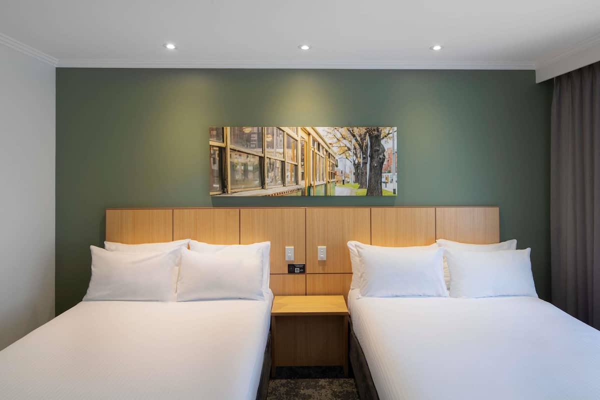 Une chambre d’hôtel avec deux lits, une tête de lit en bois, un tableau au-dessus des lits et une table de chevet entre les deux.