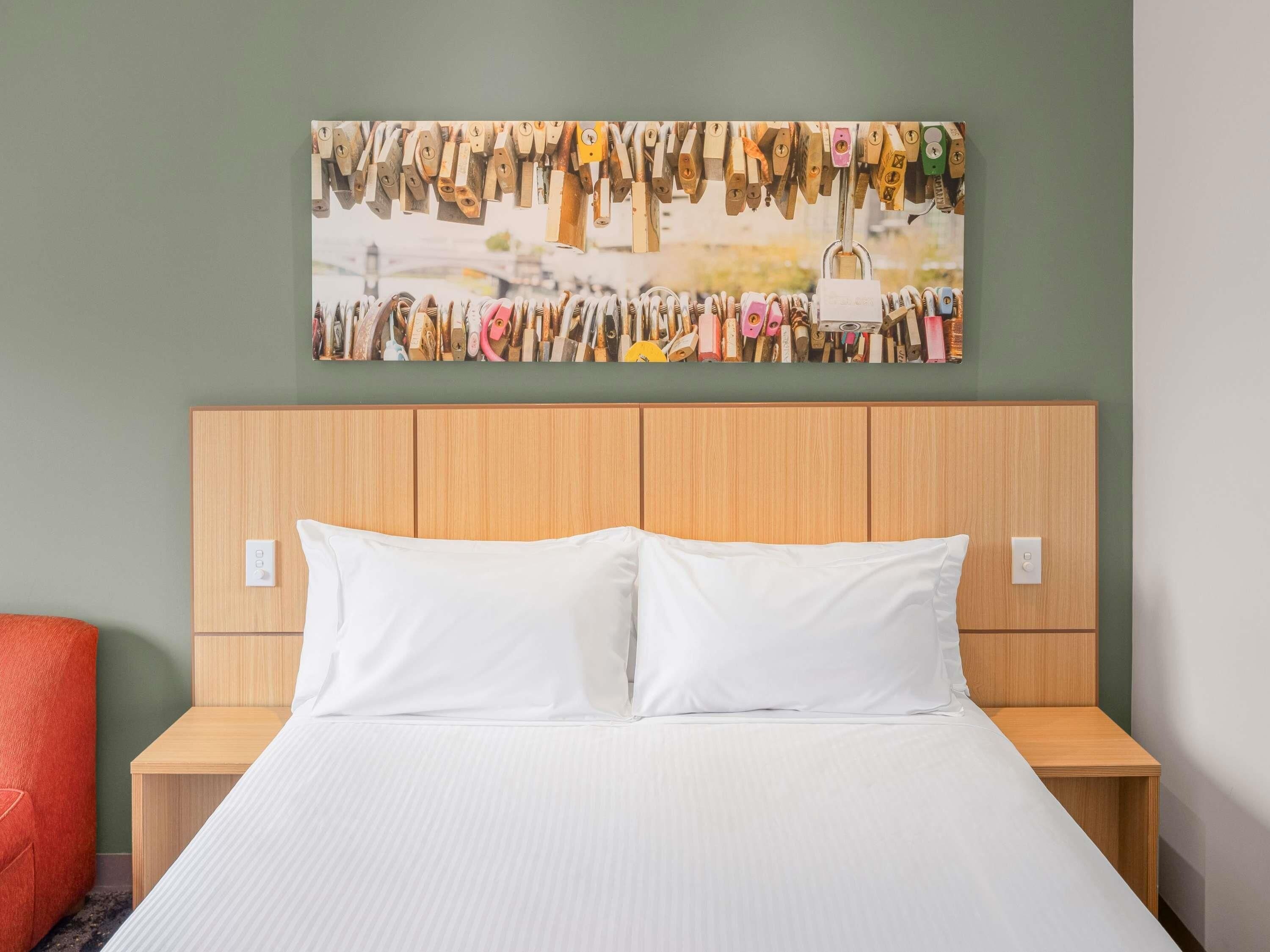 A hotel room with a wooden headboard, white bedding, and a wall-mounted artwork of various padlocks.