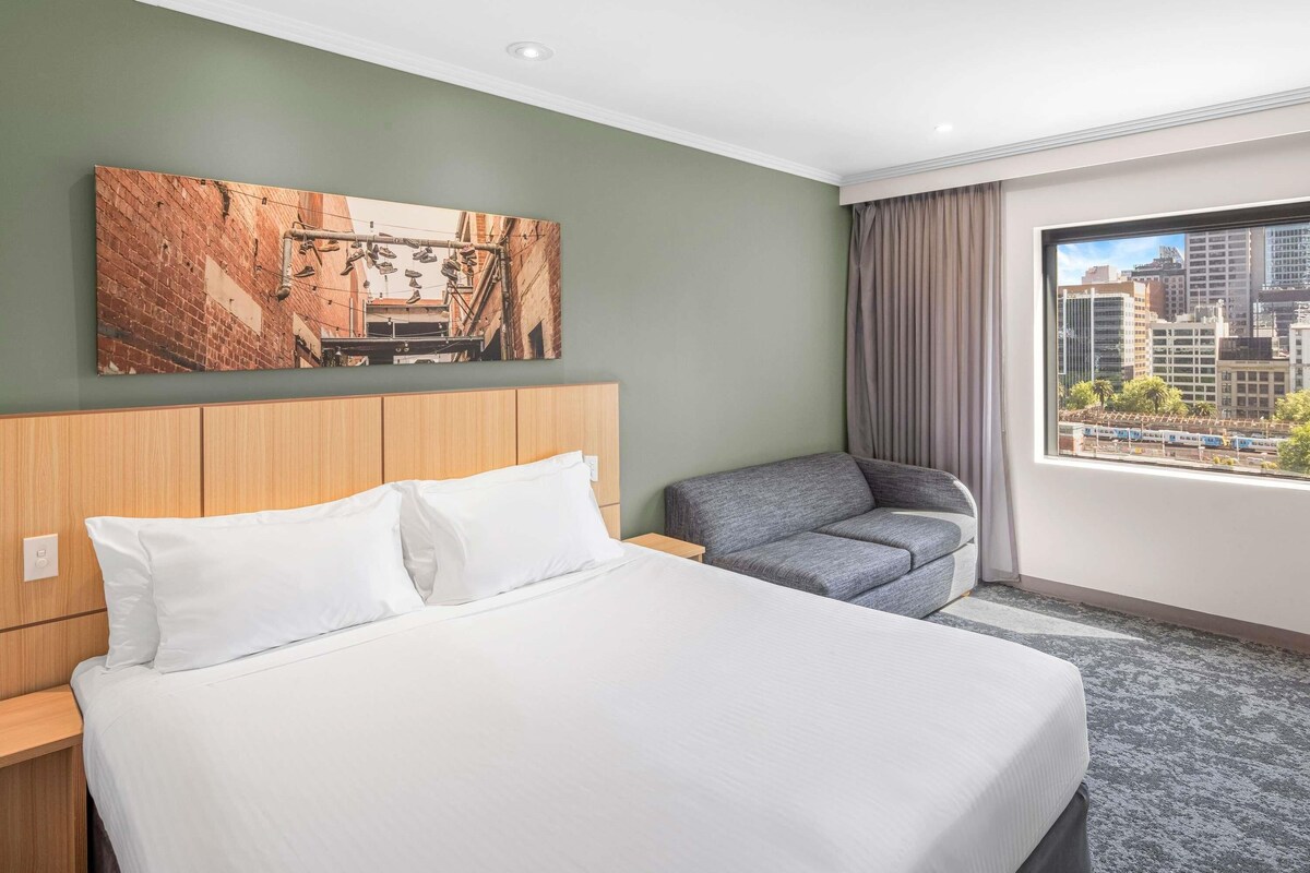 Une chambre d’hôtel avec un grand lit, un fauteuil gris, une vue sur la ville et un tableau au mur.