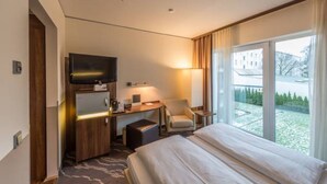 A hotel room with a bed, a desk with a computer, a chair, a TV, and a large window with a view of a building and trees.