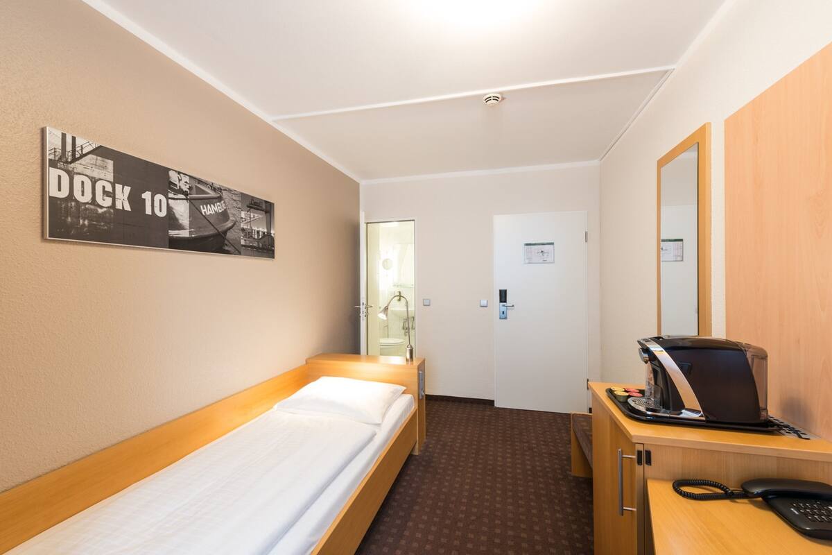 A hotel room with a single bed, a desk with a telephone, a mirror, and a poster on the wall.