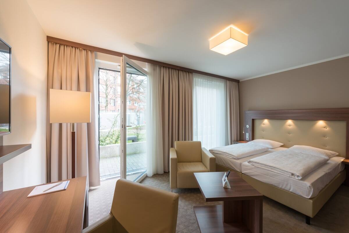 A hotel room with a bed, a desk, a chair, a lamp, and a view of the outdoors.