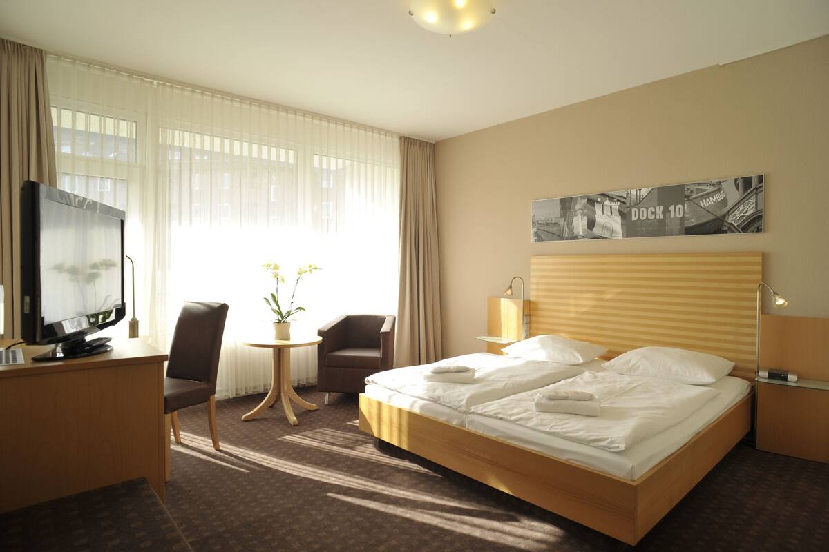 A hotel room with a bed, a desk with a TV, a chair, a small table with flowers, and a wall-mounted picture.