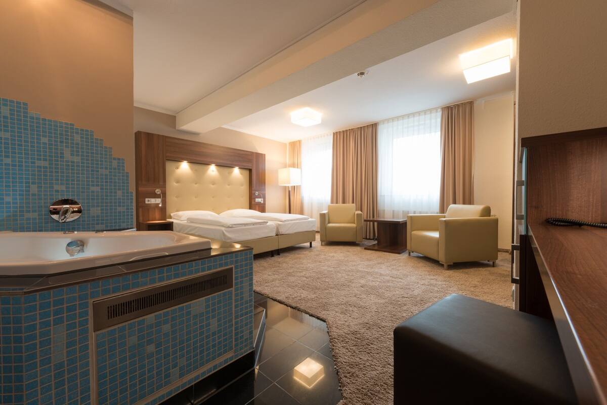 A hotel room with a large bathtub, a bed, two armchairs, a small table, and a television.