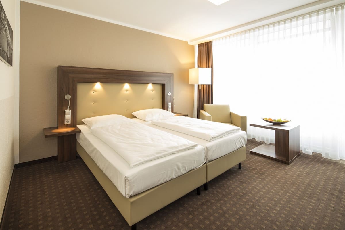 A hotel room with a double bed, a bedside table with a lamp, a small desk with a bowl of fruit, and a window with curtains.