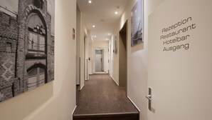 Un couloir doté d’un plancher en bois et de murs ornés de photographies en noir et blanc. Une pancarte sur le mur de droite indique : « Réception, restaurant, bar de l’hôtel, sortie ».