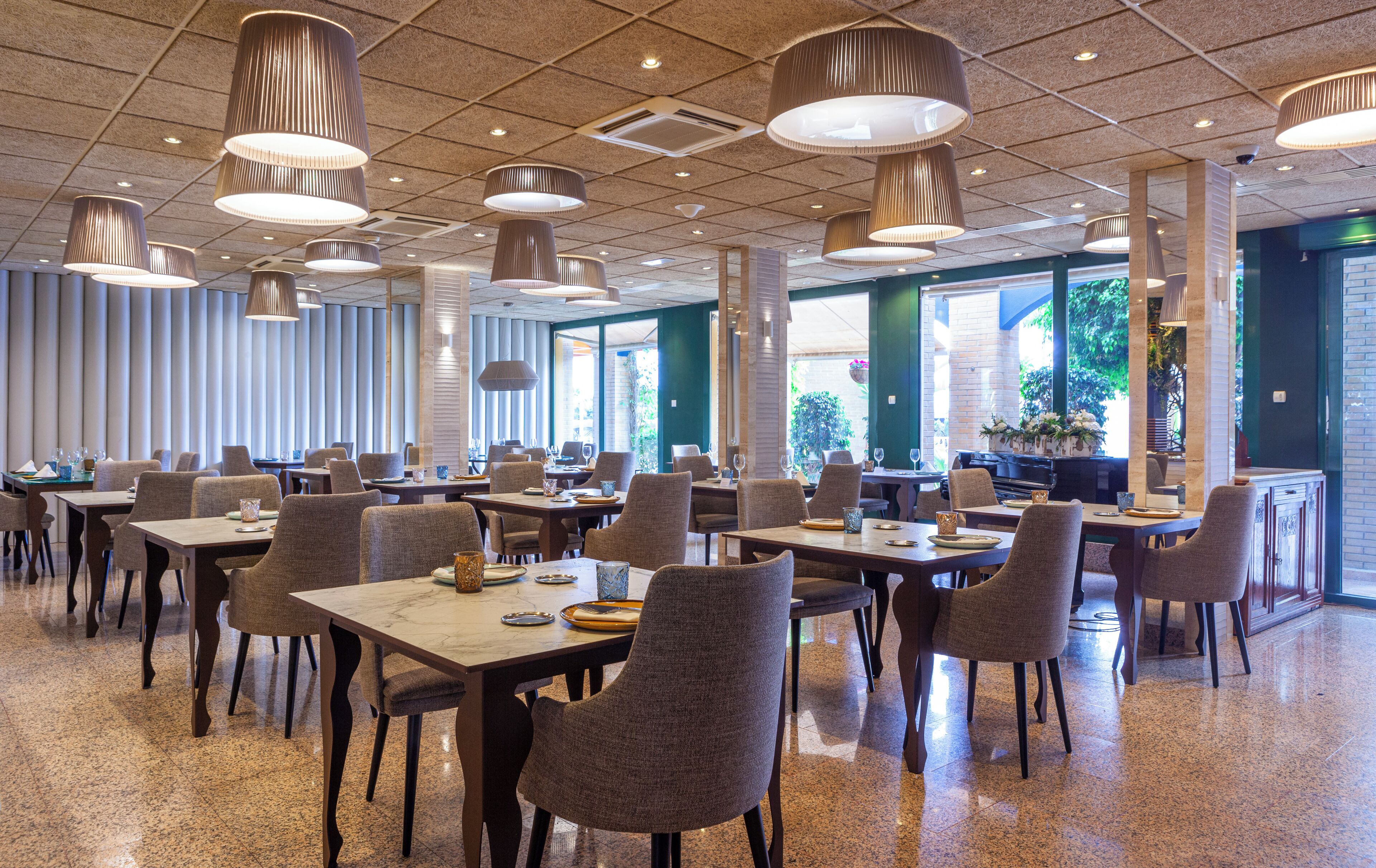 A spacious dining area with multiple tables and chairs, large hanging pendant lights, and large windows providing natural light.