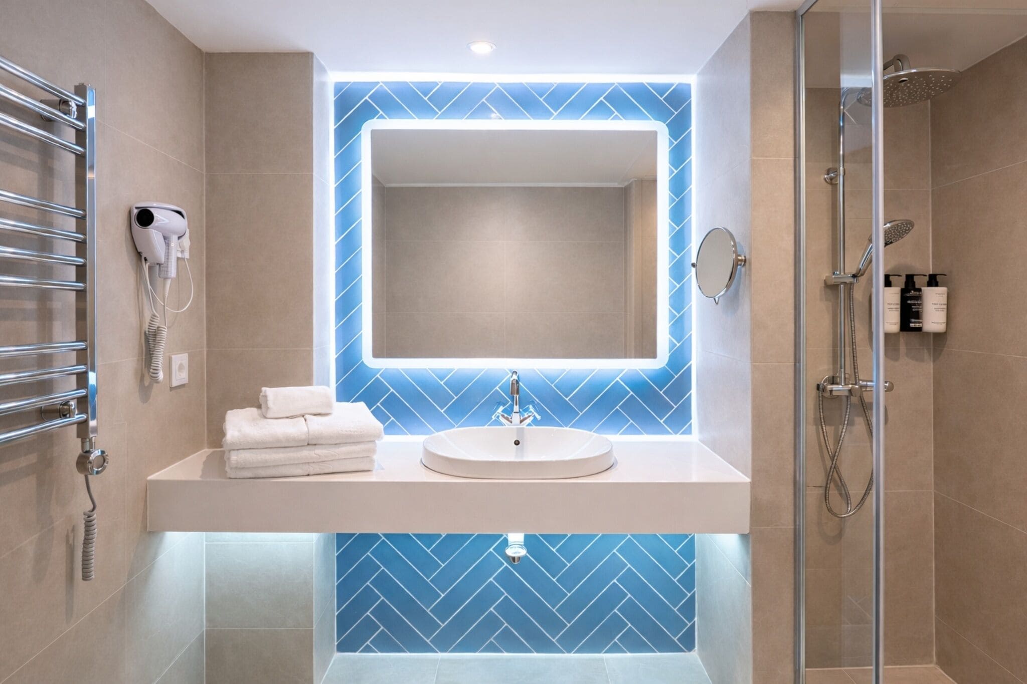 Un baño moderno con un lavamanos blanco, un espejo grande, un secador de pelo de mano y una mampara de ducha de vidrio.
