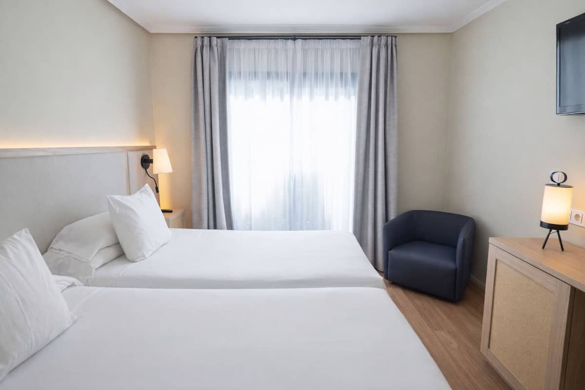 A hotel room with two beds, a desk with a lamp, a chair, and a large window with curtains.
