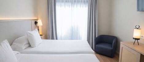 A hotel room with two beds, a desk with a lamp, a chair, and a large window with curtains.