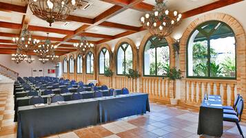 Un salón de conferencias con filas de mesas y sillas, ventanas amplias y varios candelabros.