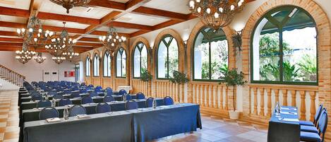 Un salón de conferencias con filas de mesas y sillas, ventanas amplias y varios candelabros.