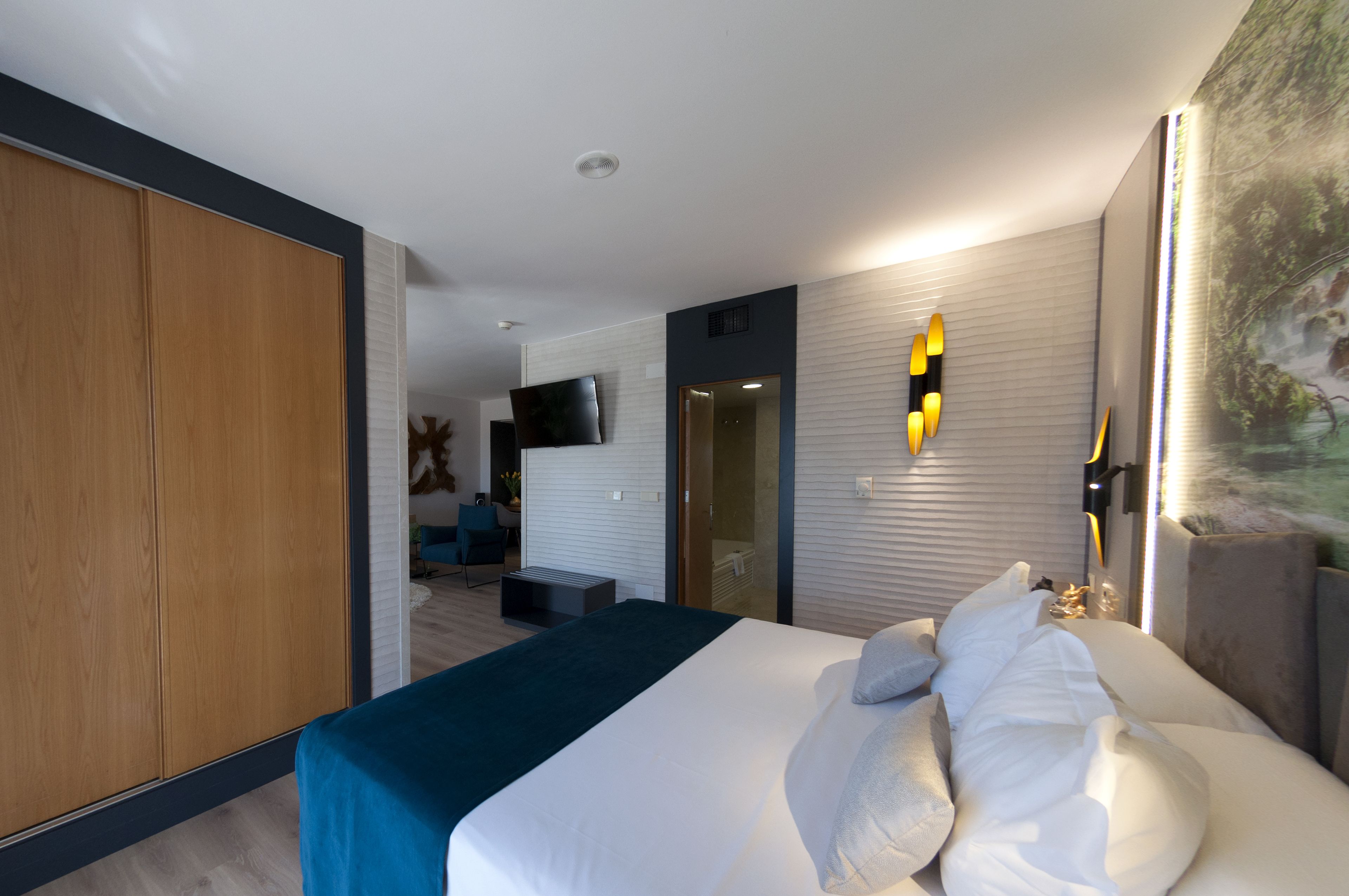 A modern hotel room with a large bed, a wooden wardrobe, a seating area with a chair and a small table, a flat-screen TV mounted on the wall, and a large window with blinds.