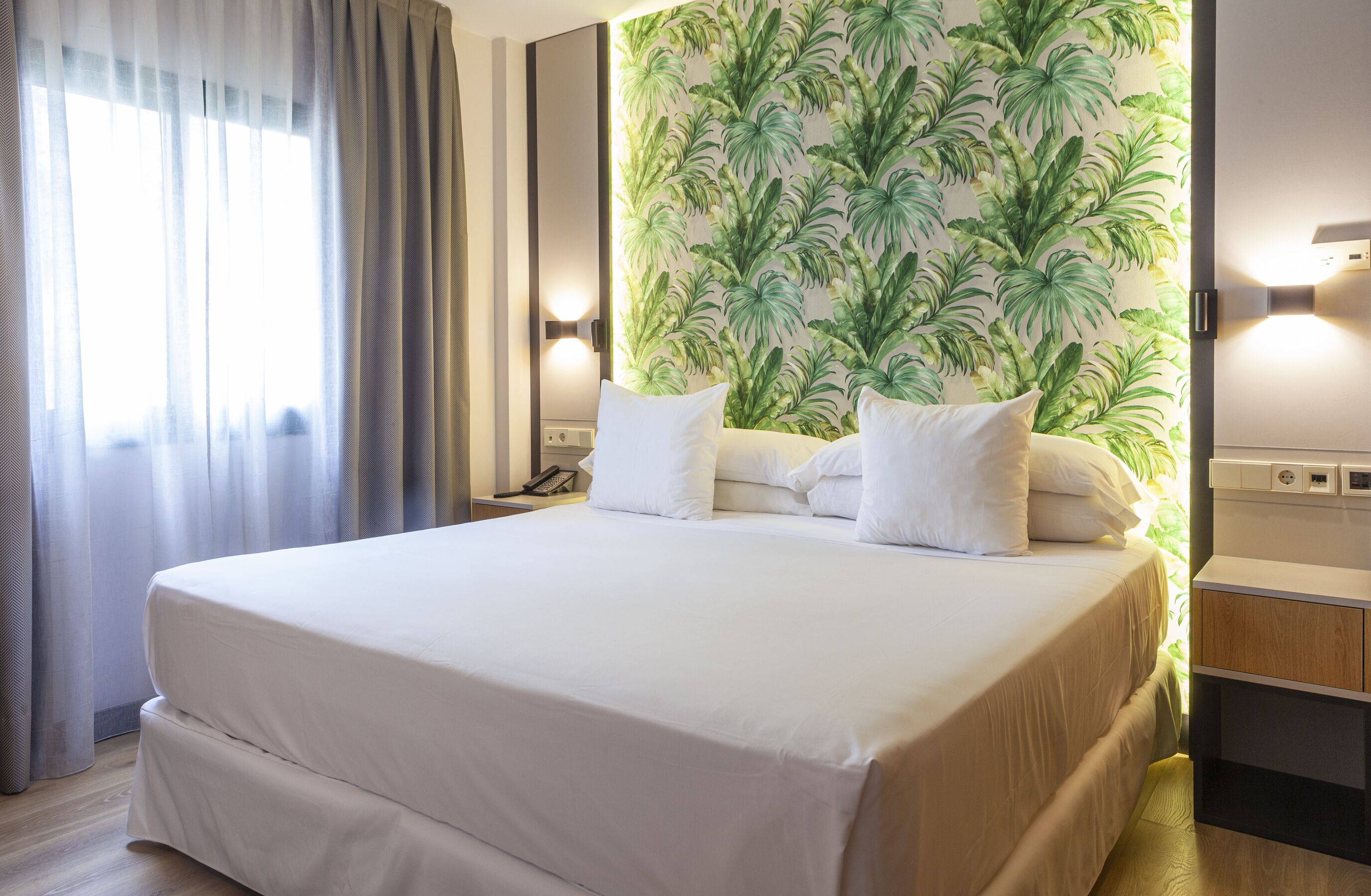 Ein Hotelzimmer mit einem großen Bett, einem Nachttisch mit Telefon, einer Wand mit tropischem Blattmuster und einem Fenster mit Vorhängen.