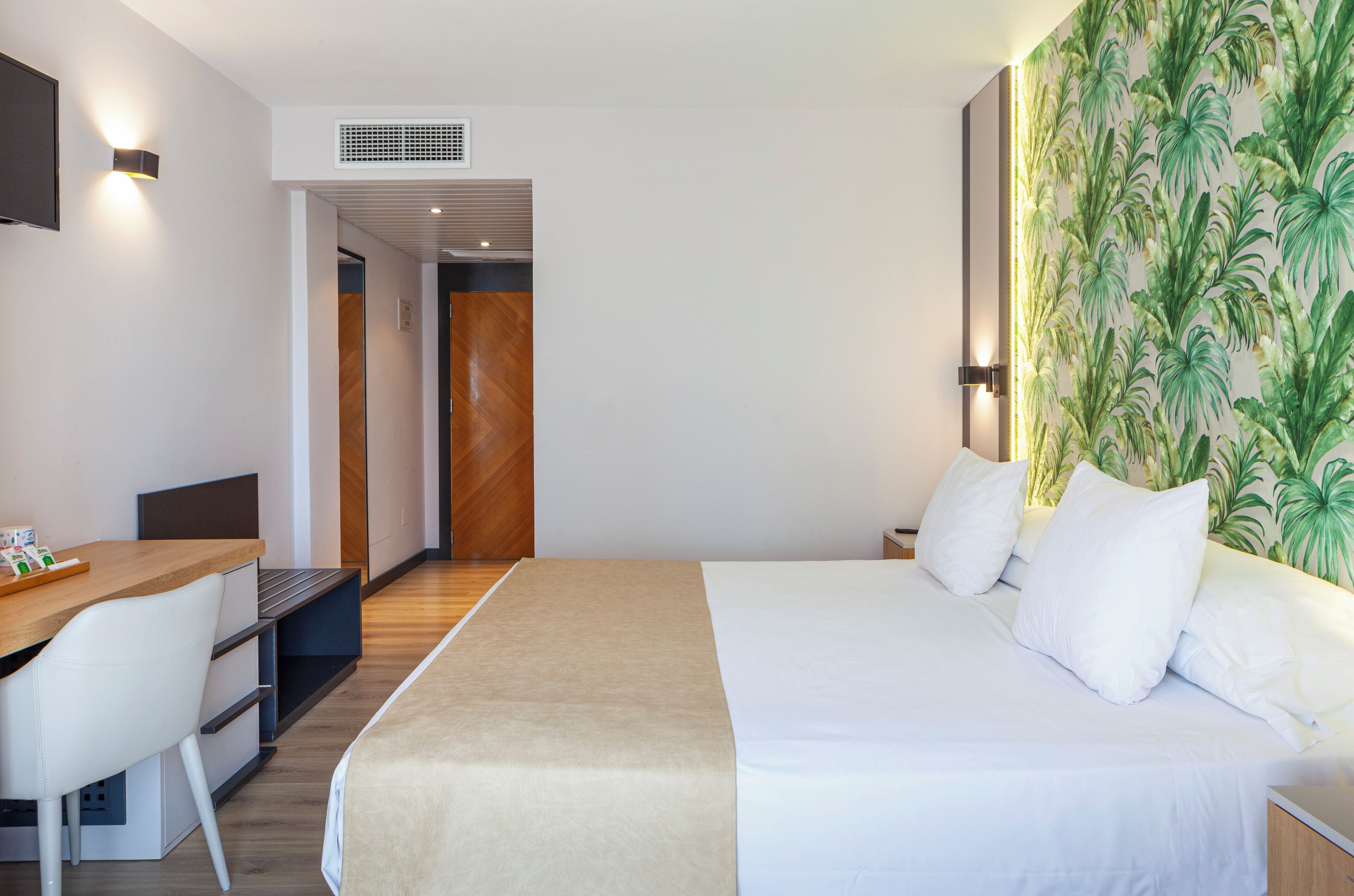 Habitación de hotel con cama, escritorio y silla. Hay aire acondicionado de pared y una pared con papel tapiz tropical.