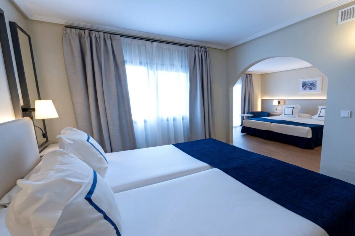 A hotel room with two beds, a sitting area, and a view of the adjacent room.