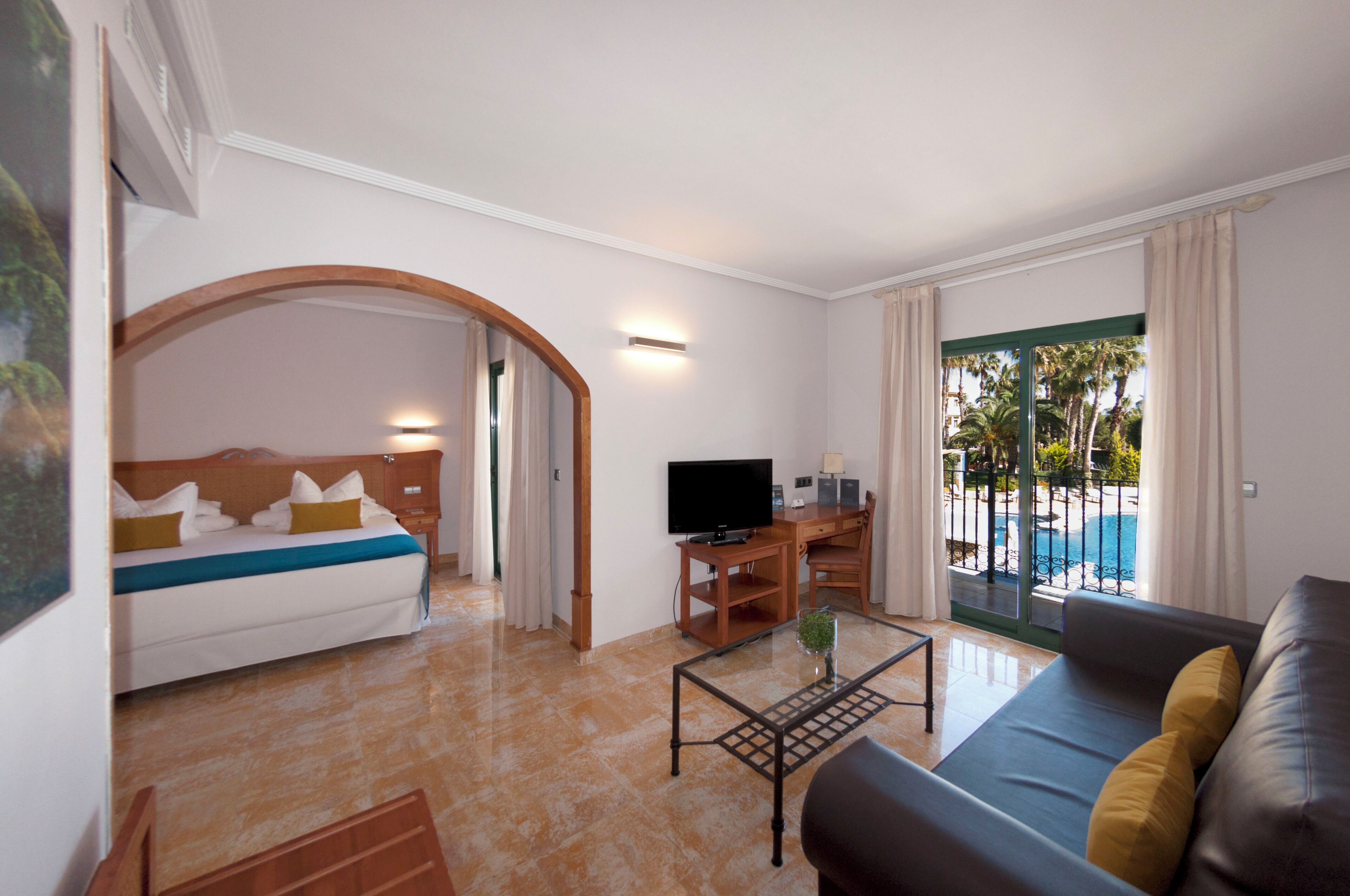 A hotel room with a bed, a TV, a sofa, a coffee table, and a view of a pool.