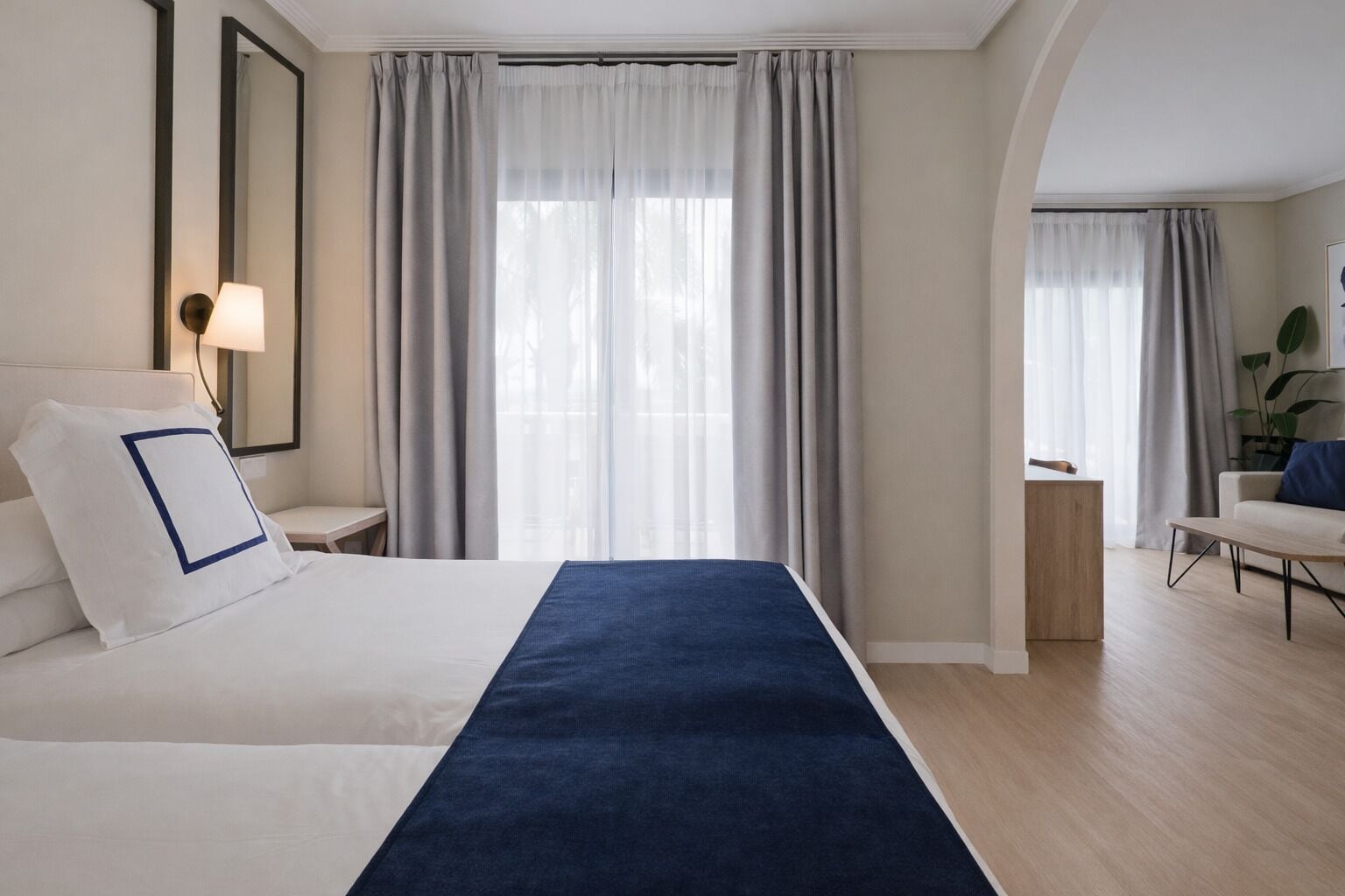 Una habitación de hotel moderna con una cama grande, una mesita de noche con lámpara, una pequeña mesita auxiliar de madera y una zona de estar con una silla y una mesa.