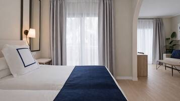 Una habitación de hotel moderna con una cama grande, una mesita de noche con lámpara, una pequeña mesita auxiliar de madera y una zona de estar con una silla y una mesa.