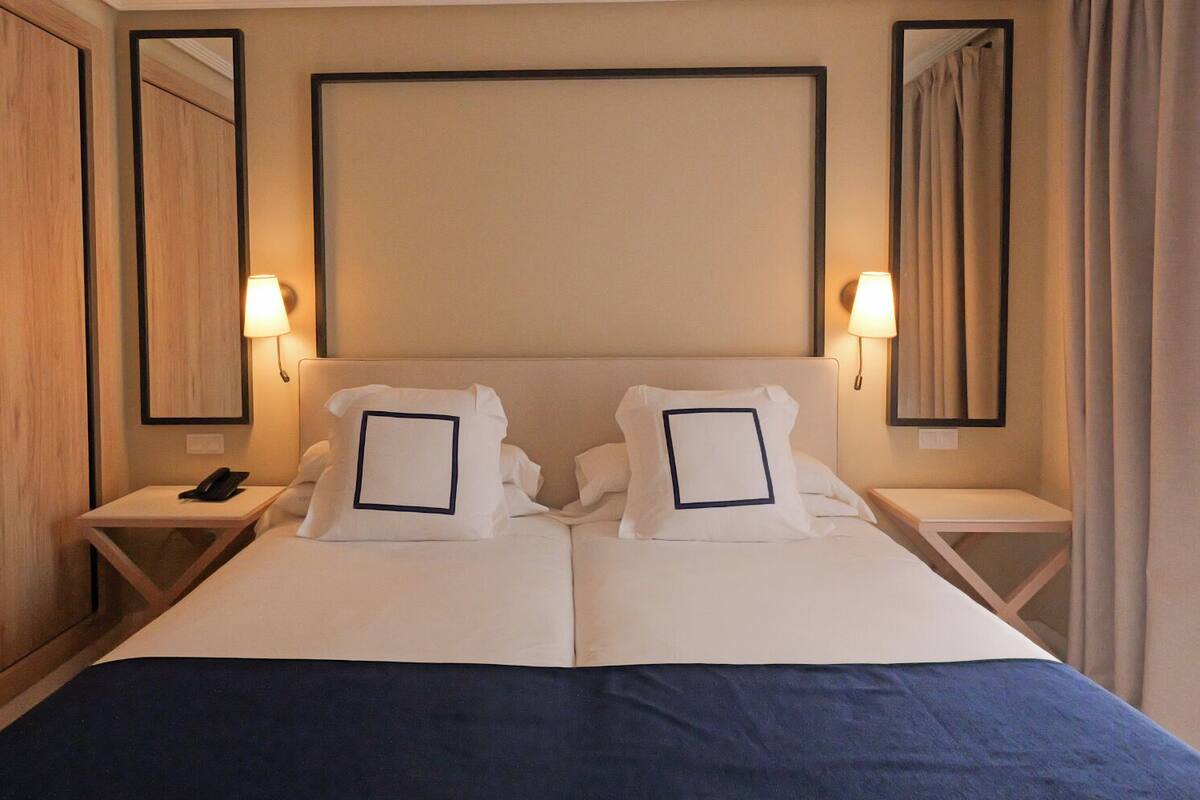 A double bed with a blue bedspread, two white pillows with black borders, two bedside tables with lamps, and a wooden headboard.
