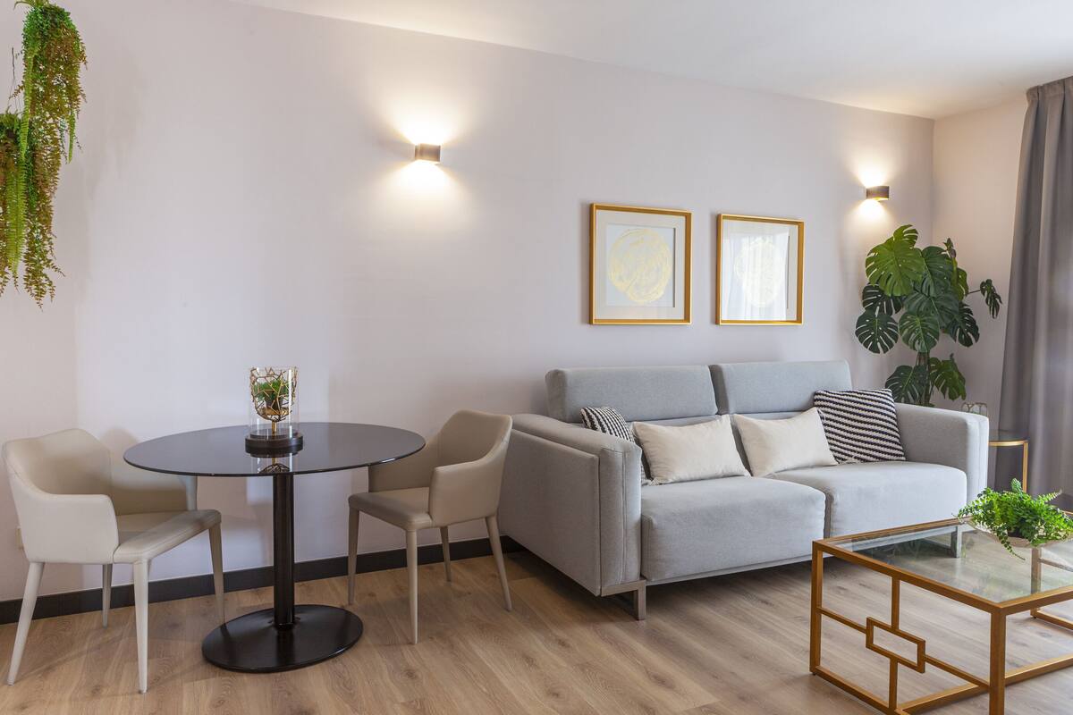A modern living room with a grey sofa, two armchairs, a round glass table, and a coffee table with a geometric design. There are two framed artworks on the wall, a potted plant, and a hanging plant.