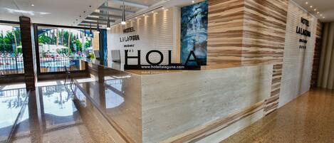 A modern hotel reception area with a glass wall offering a view of a pool and outdoor seating.