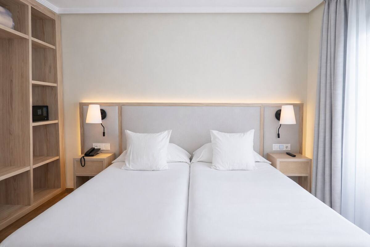A double bed with white linens, two bedside tables with lamps, a wooden wardrobe, and a neutral-colored wall.