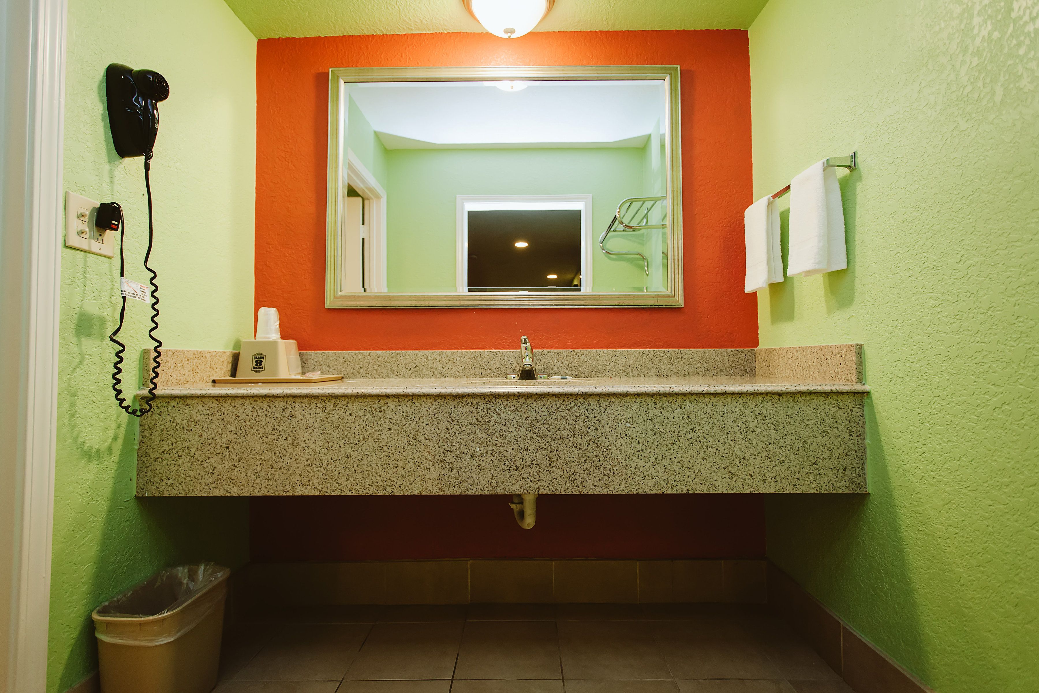 A bathroom with a granite countertop sink, a mirror, a hairdryer, and a trash bin.