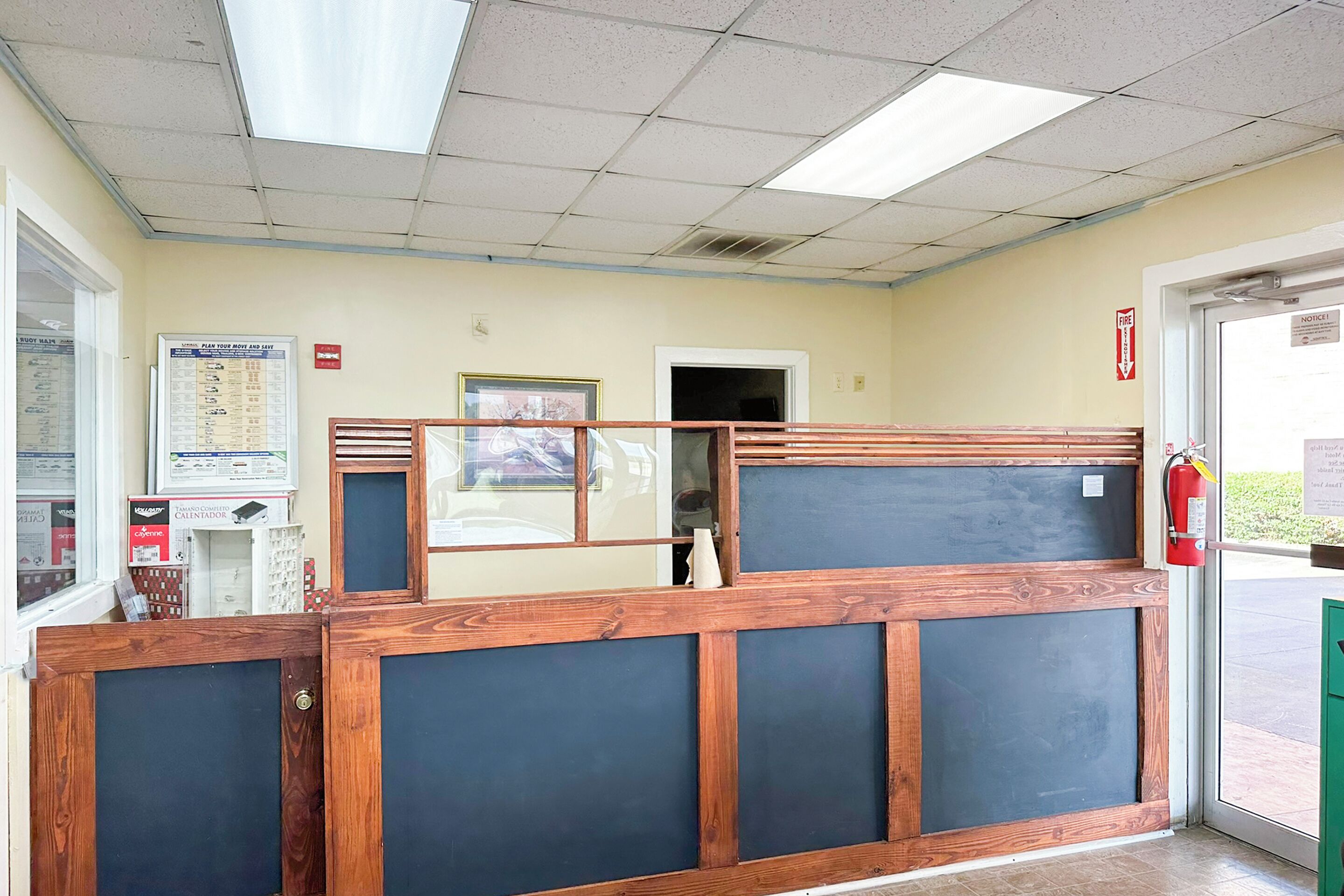 Une salle d’attente avec un bureau en bois, un extincteur, un tableau d’affichage et une porte donnant sur l’extérieur.