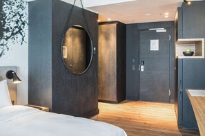 A modern hotel room with a large bed, a circular mirror, a wooden floor, a door with a sign, and a small shelf with a bowl.