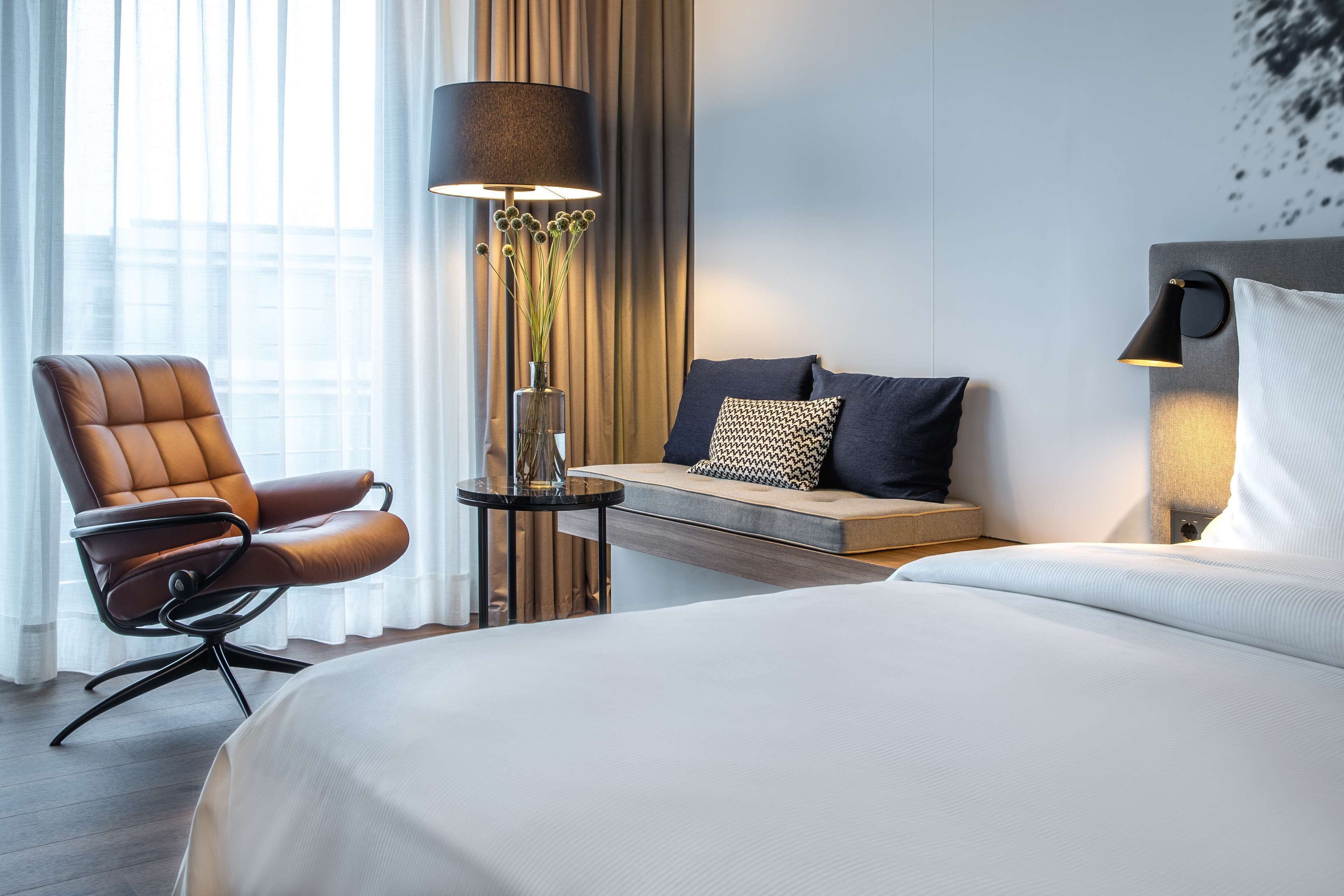 Ett hotellrum med en stor säng, en brun läderfåtölj, ett litet bord med en lampa, en bänk med kuddar och en vägglampa.