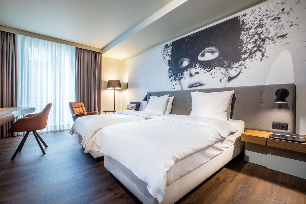 Ein Hotelzimmer mit zwei Betten, einem Schreibtisch, einem Stuhl, einer Lampe und einem großen Wandbild.