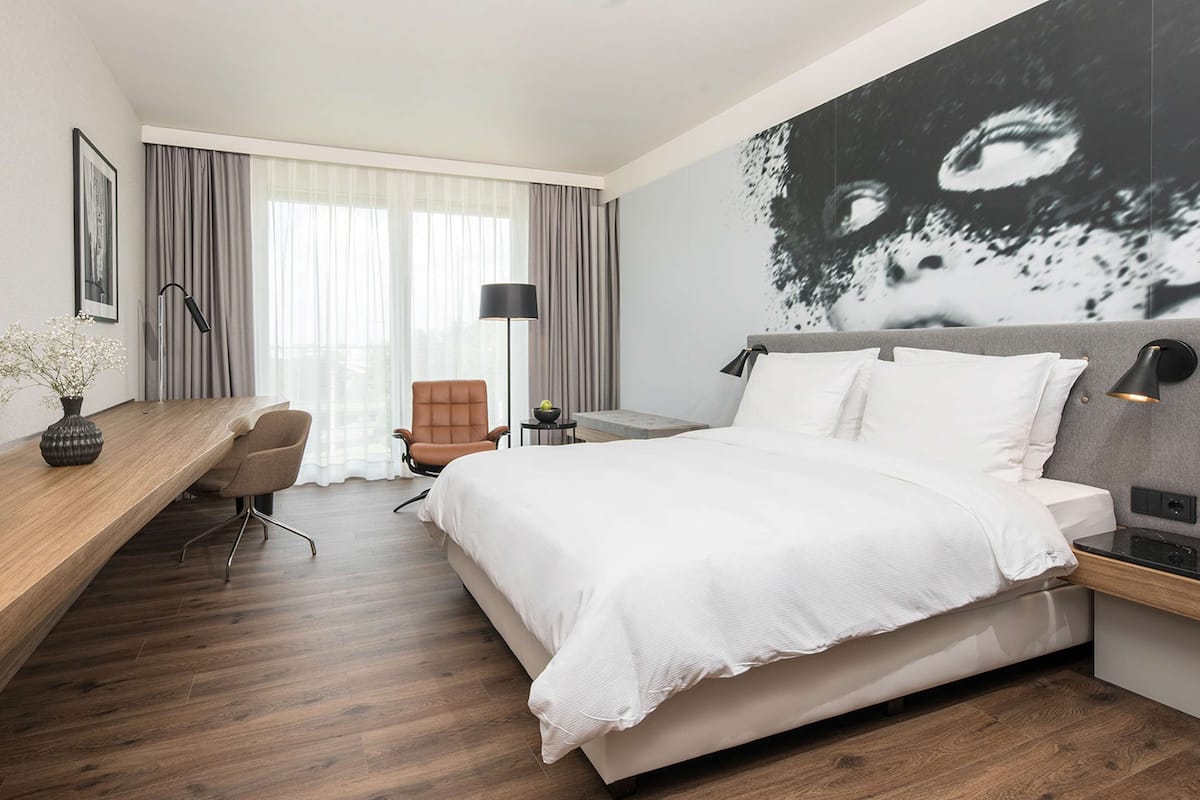 Ein modernes Hotelzimmer mit einem großen Bett, einem Schreibtisch mit Stuhl, einem kleinen Tisch und einer Wandlampe.
