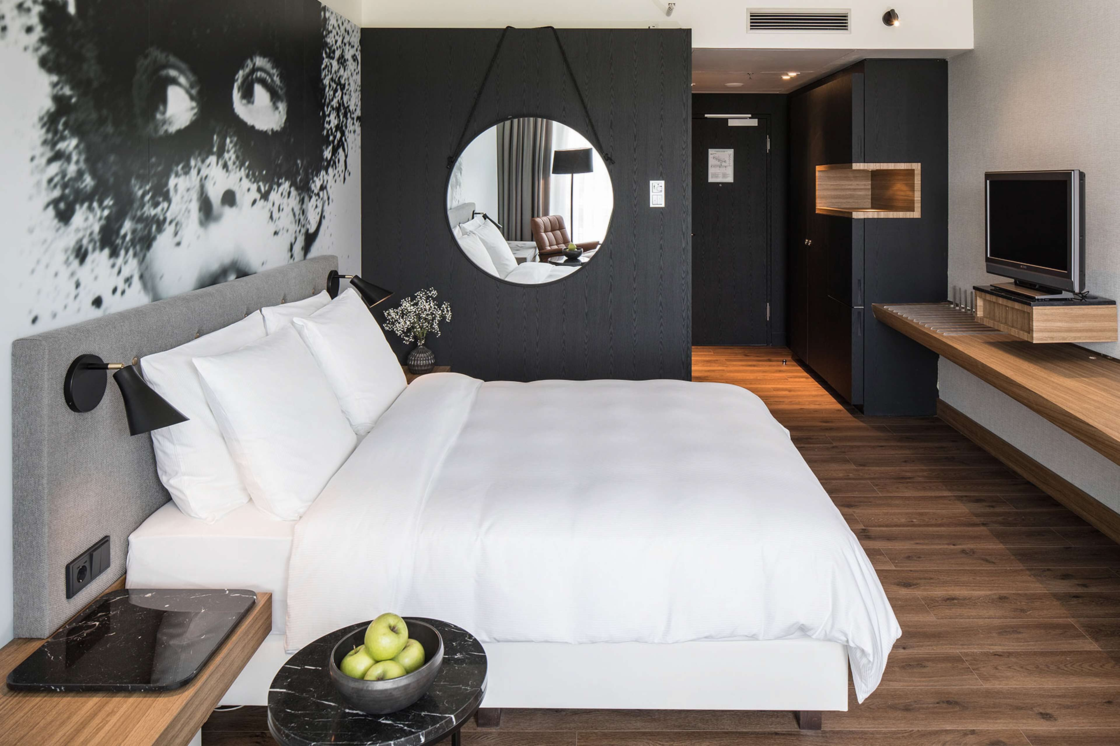 A modern hotel room with a large bed, a round mirror, a TV, and a wooden side table.