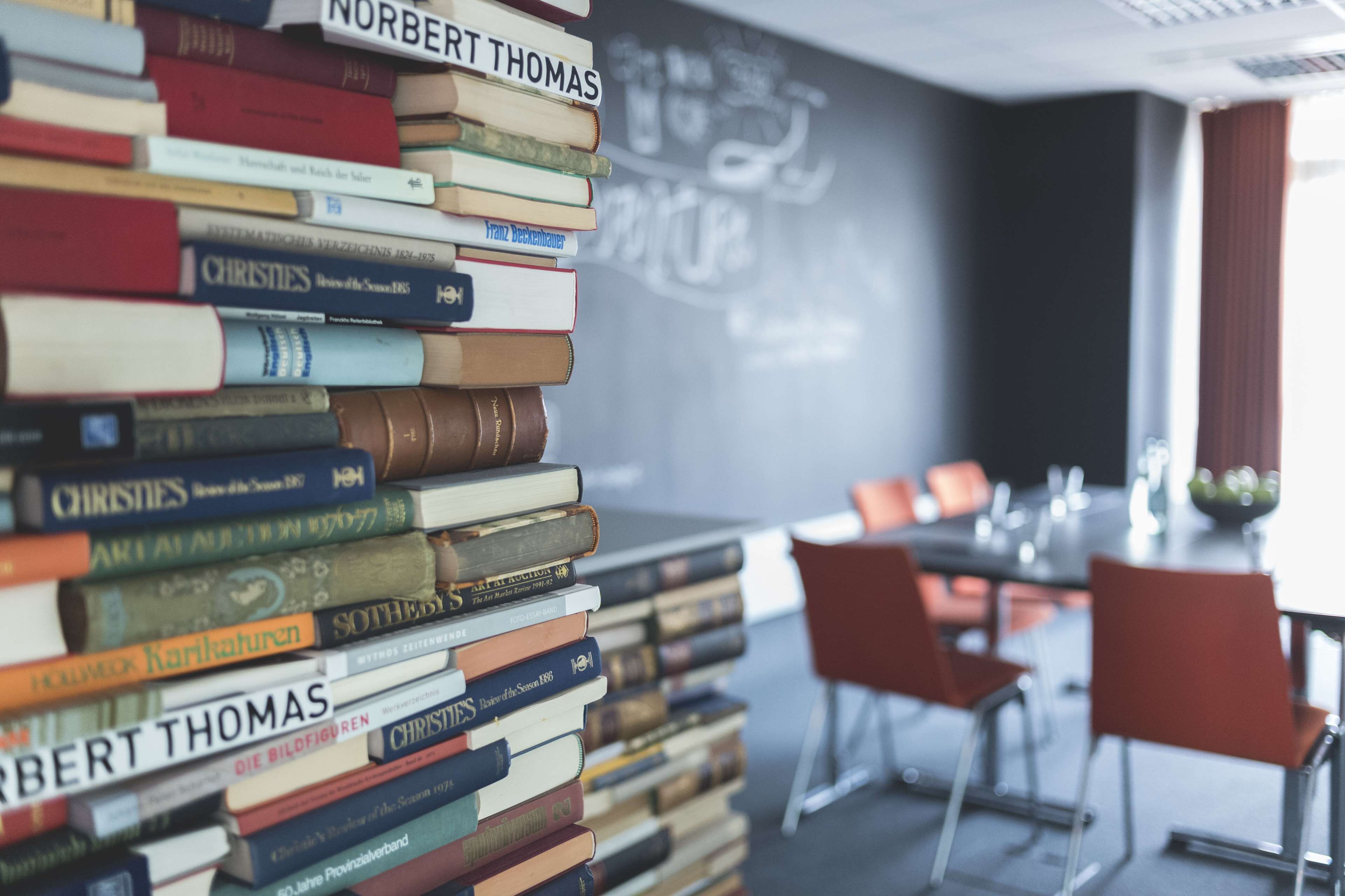 A stack of books in a room with a chalkboard wall and a dining area with chairs.