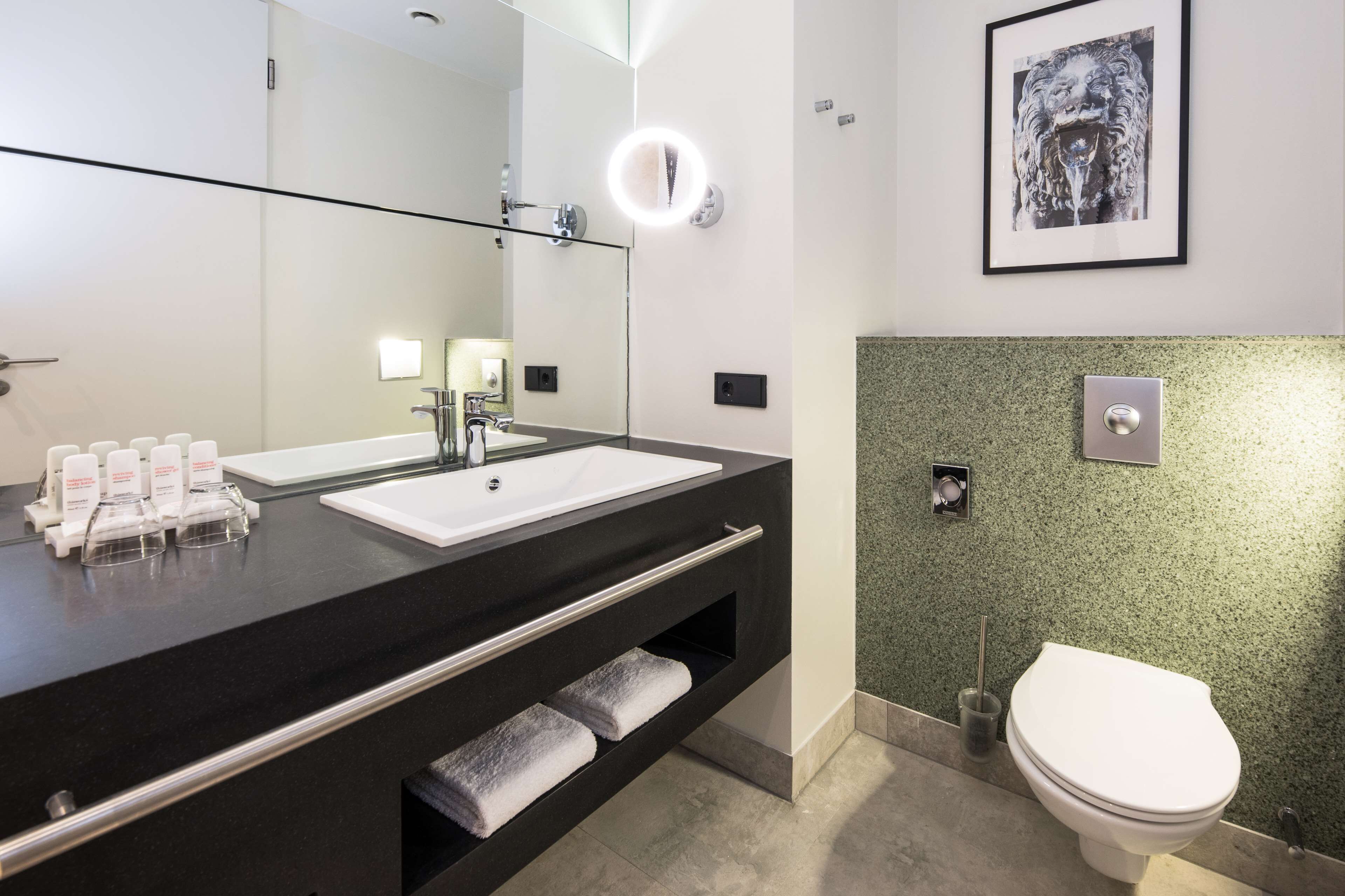 Un baño moderno con encimera negra, lavabo blanco y un espejo grande con marco iluminado. Hay un inodoro con pared de aspecto moteado verde y una foto enmarcada encima.