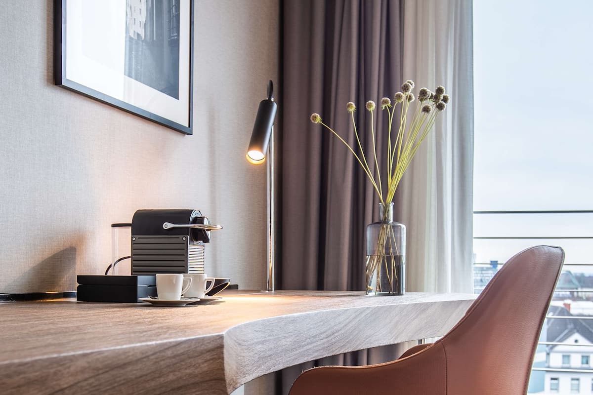 Ein modernes Hotelzimmer mit Schreibtisch aus Holz, einer Kaffeemaschine, einer Vase mit Trockenblumen und Blick auf Gebäude durch das Fenster.