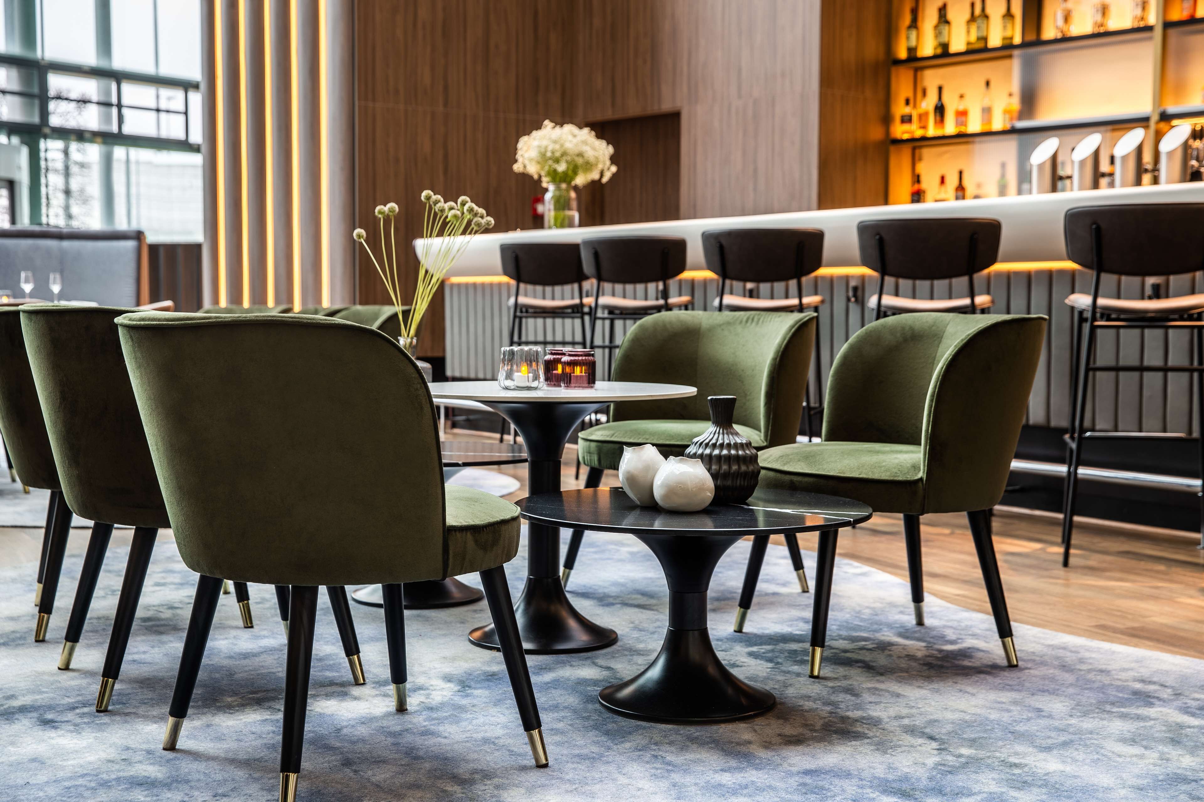 A modern bar area with green upholstered chairs, a round table, and a bar counter with bottles.