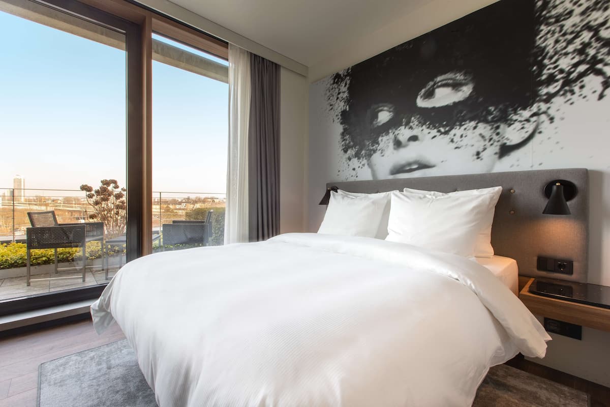 Ein modernes Hotelzimmer mit einem großen Bett, Blick auf die Stadt durch raumhohe Fenster und einem großflächigen Wandbild.
