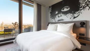 Une chambre d’hôtel moderne dotée d’un grand lit, offrant une vue sur la ville grâce à de larges fenêtres du sol au plafond, et agrémentée d’une grande fresque murale.
