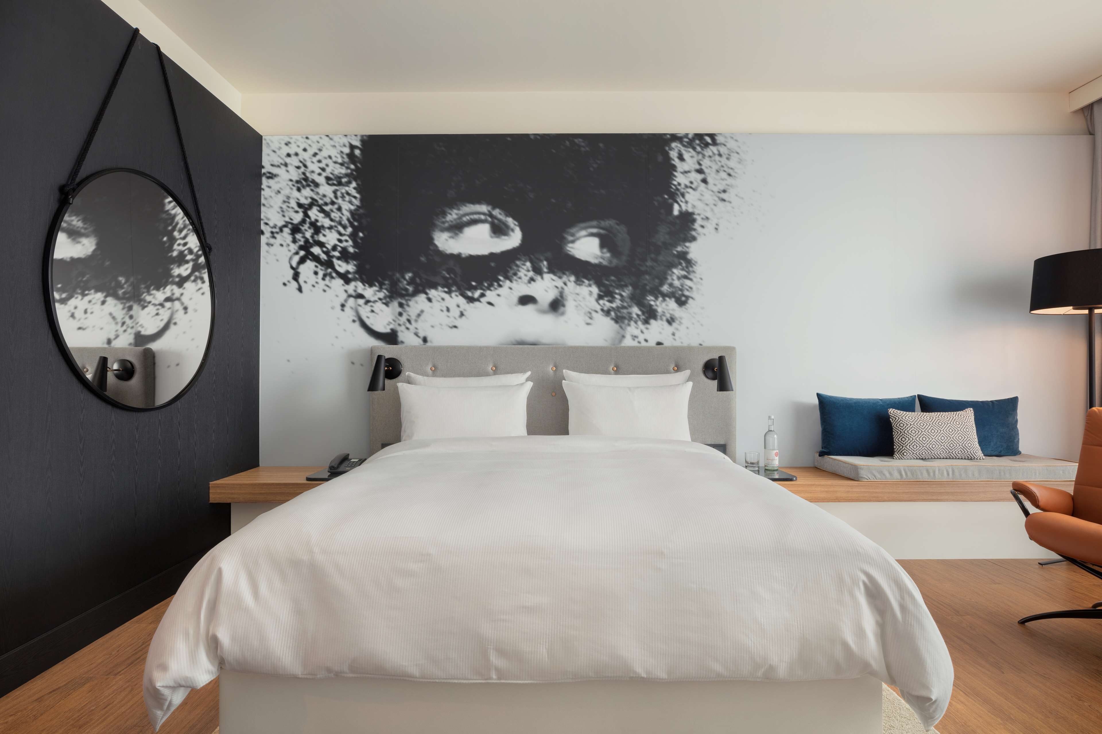 A modern hotel room with a large bed, a bedside table with a lamp, a wall-mounted mirror, and a large abstract artwork.