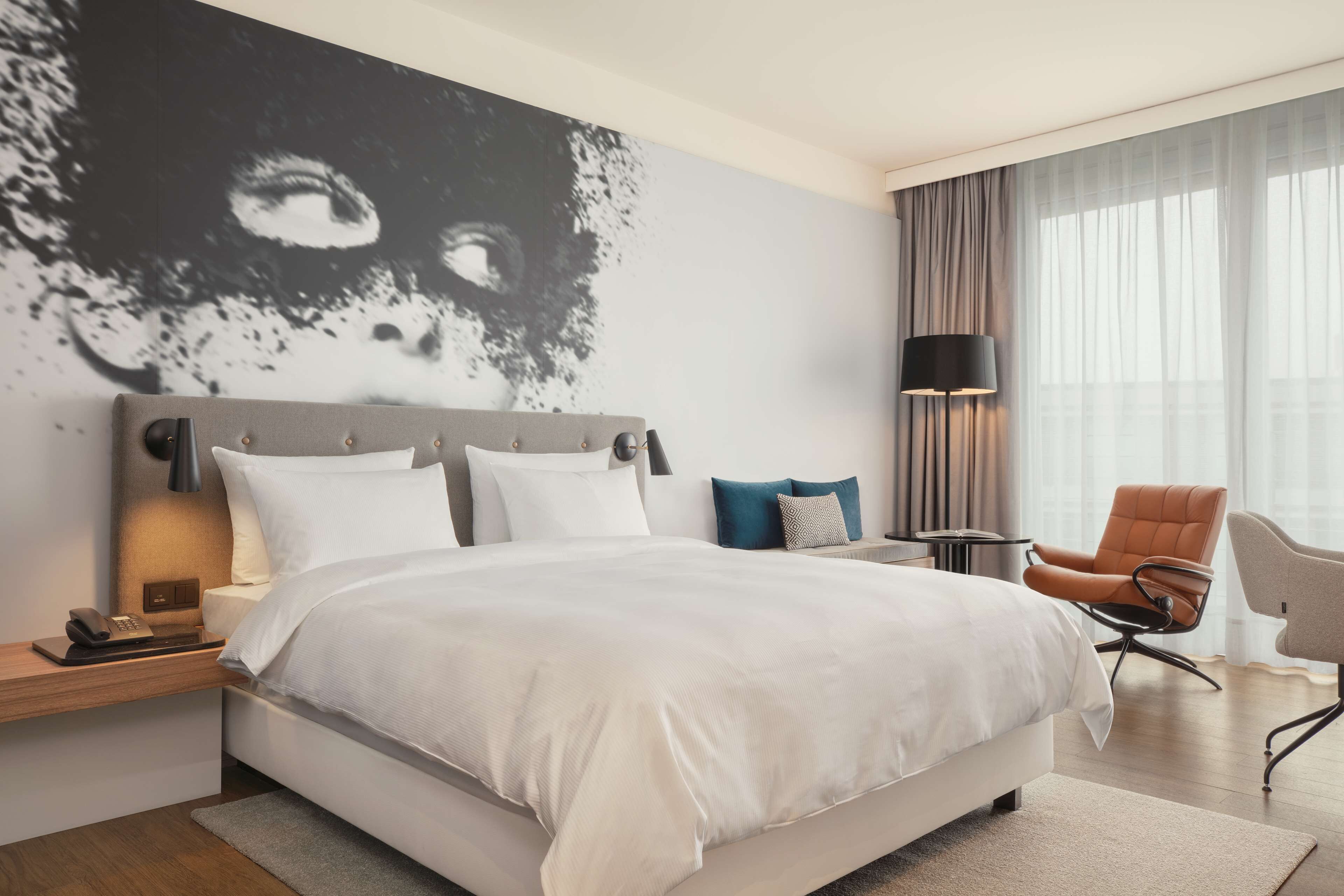 Ein modernes Hotelzimmer mit einem großen Bett, einem Nachttisch mit Lampe, einem Sessel und einem großen abstrakten Gemälde an der Wand.
