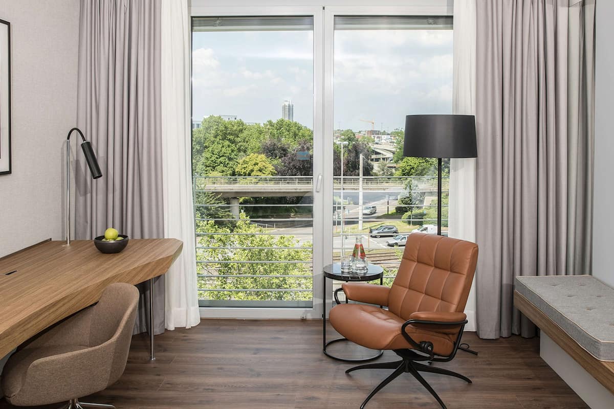 Ein modernes Zimmer mit einem Holztisch, einem Ledersessel, einem kleinen runden Tisch mit einer Lampe und Blick auf eine Stadtlandschaft durch große Fenster.