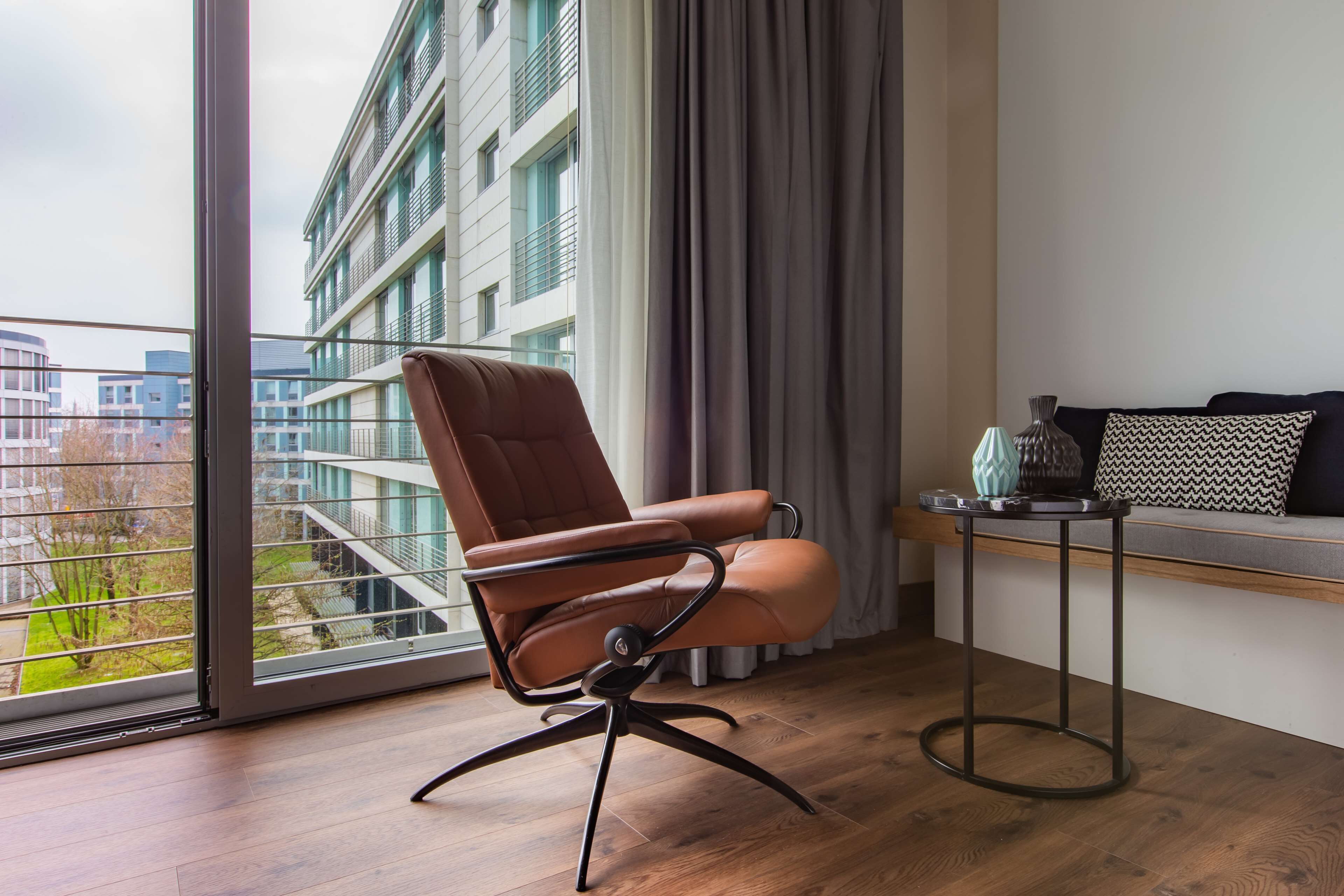 Una habitación moderna con un sillón de cuero, una mesita auxiliar y vistas a un edificio a través de un ventanal grande.