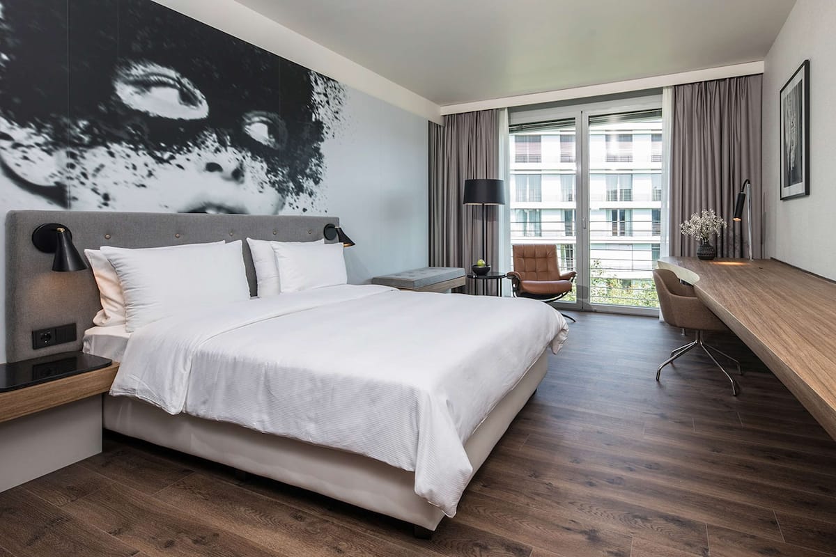 Ein modernes Hotelzimmer mit einem großen Bett, einem Schreibtisch und Blick auf die Stadt durch große Fenster.