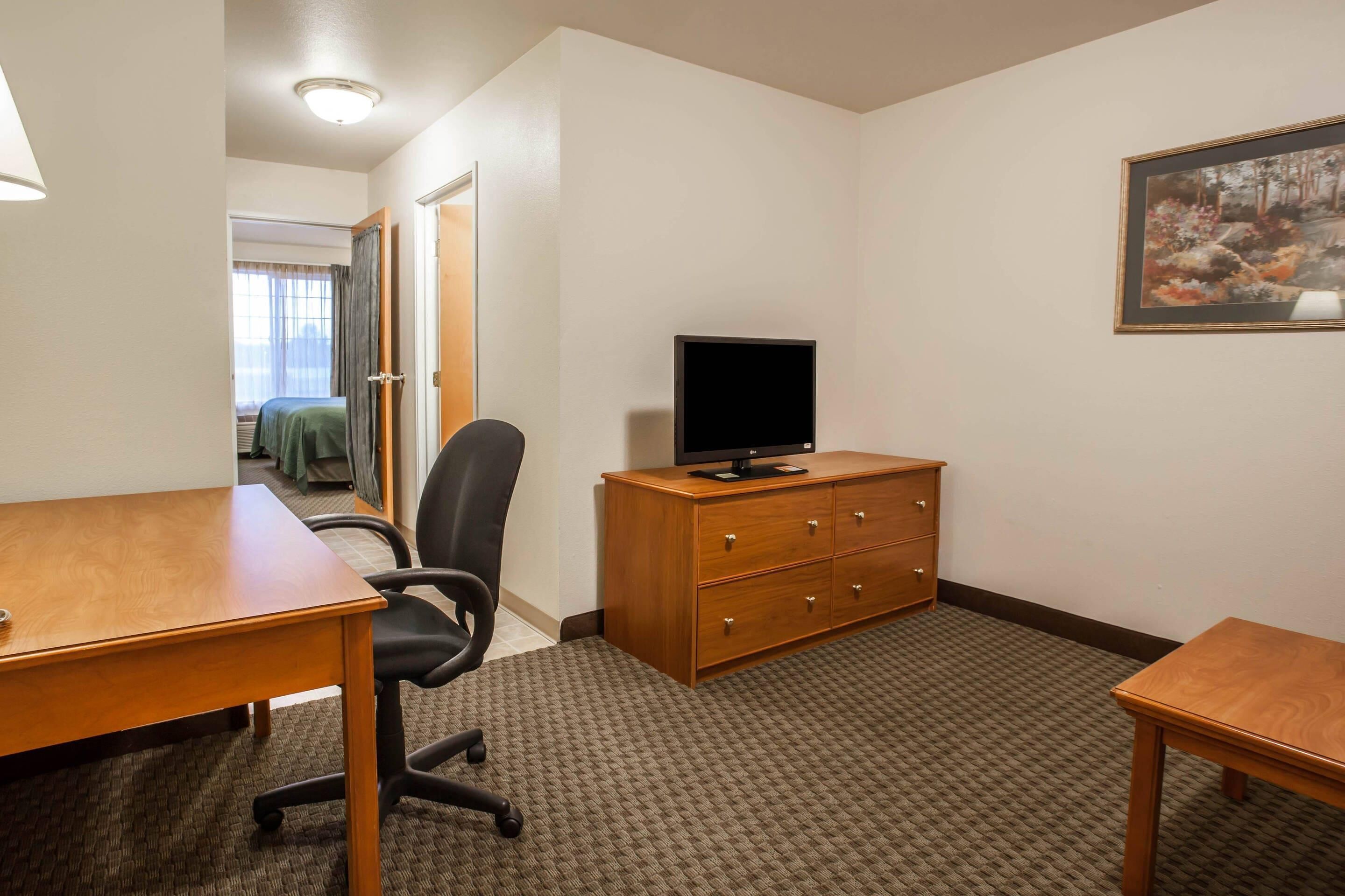A hotel room with a wooden desk, a black office chair, a wooden dresser, and a flat-screen TV.