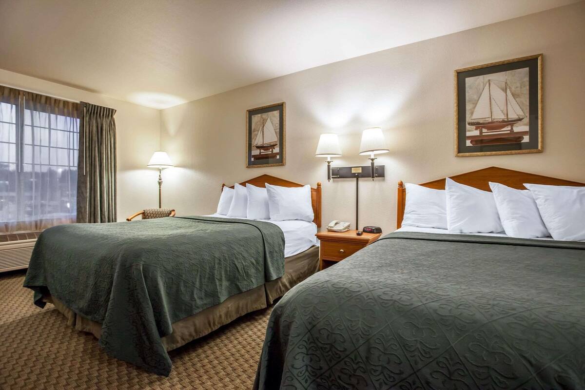 A hotel room with two beds, a nightstand with a lamp, a window with curtains, and a framed picture on the wall.