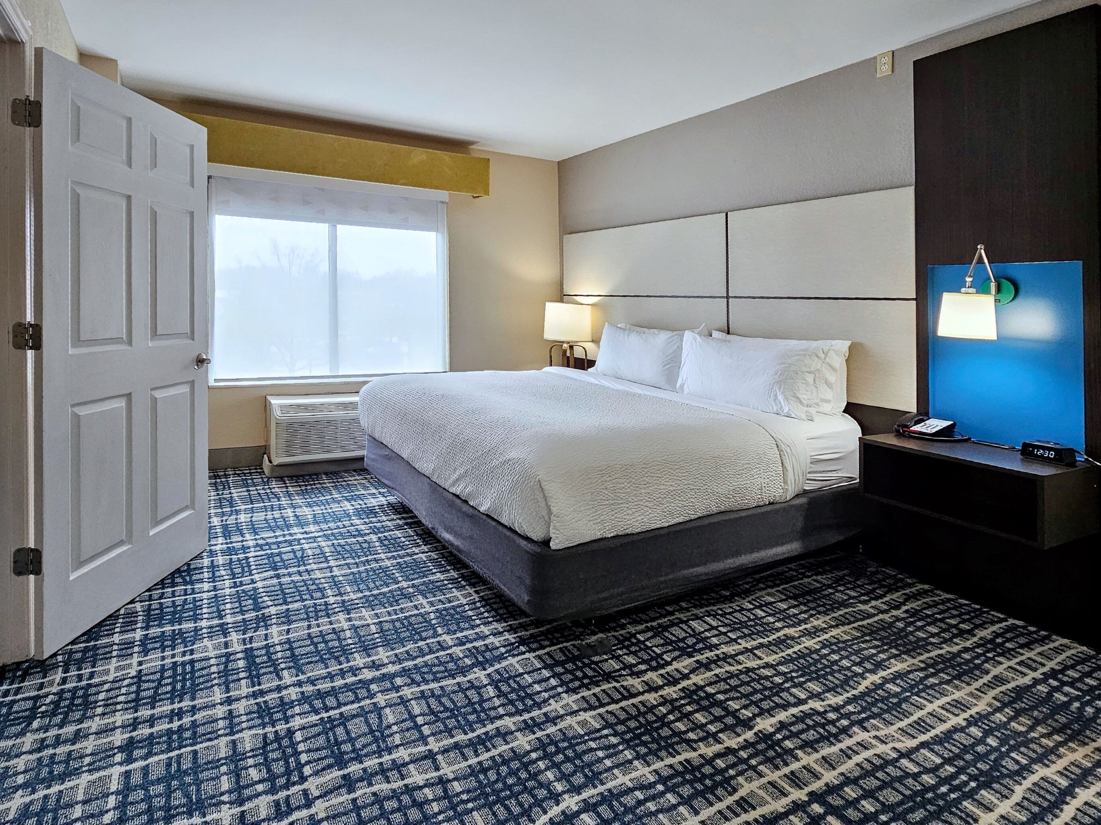 A hotel room with a large bed, a bedside table with a lamp, a nightstand with a phone, and a window with a view.
