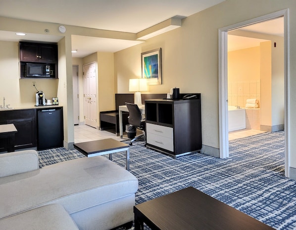 Une chambre d’hôtel équipée d’une kitchenette, d’un bureau, d’une chaise, d’un canapé et d’une salle de bain visible à travers une porte ouverte.
